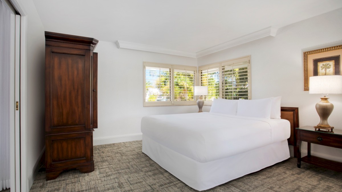 Bright room with a bed, wooden wardrobe, side tables with lamps, carpet, and windows with blinds.