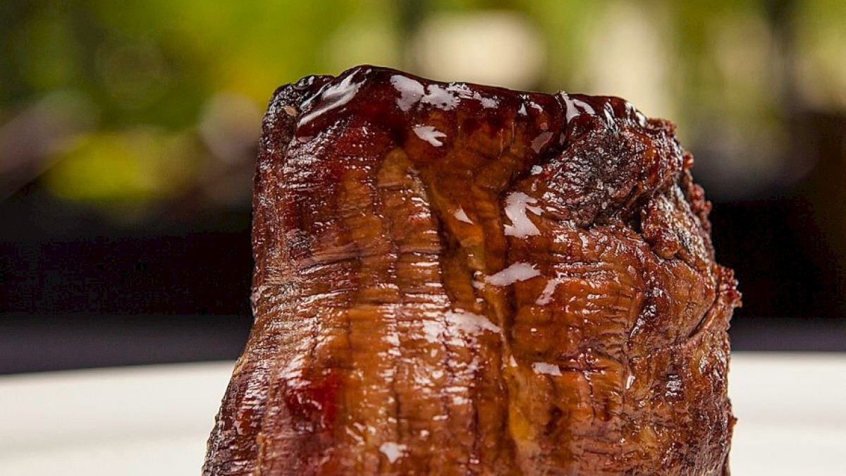 A perfectly cooked piece of steak sits on a white plate, with a blurred natural background, ending the sentence.