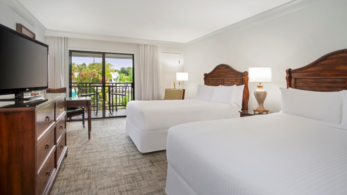 A hotel room with two beds, a desk, TV, and a balcony with a view.