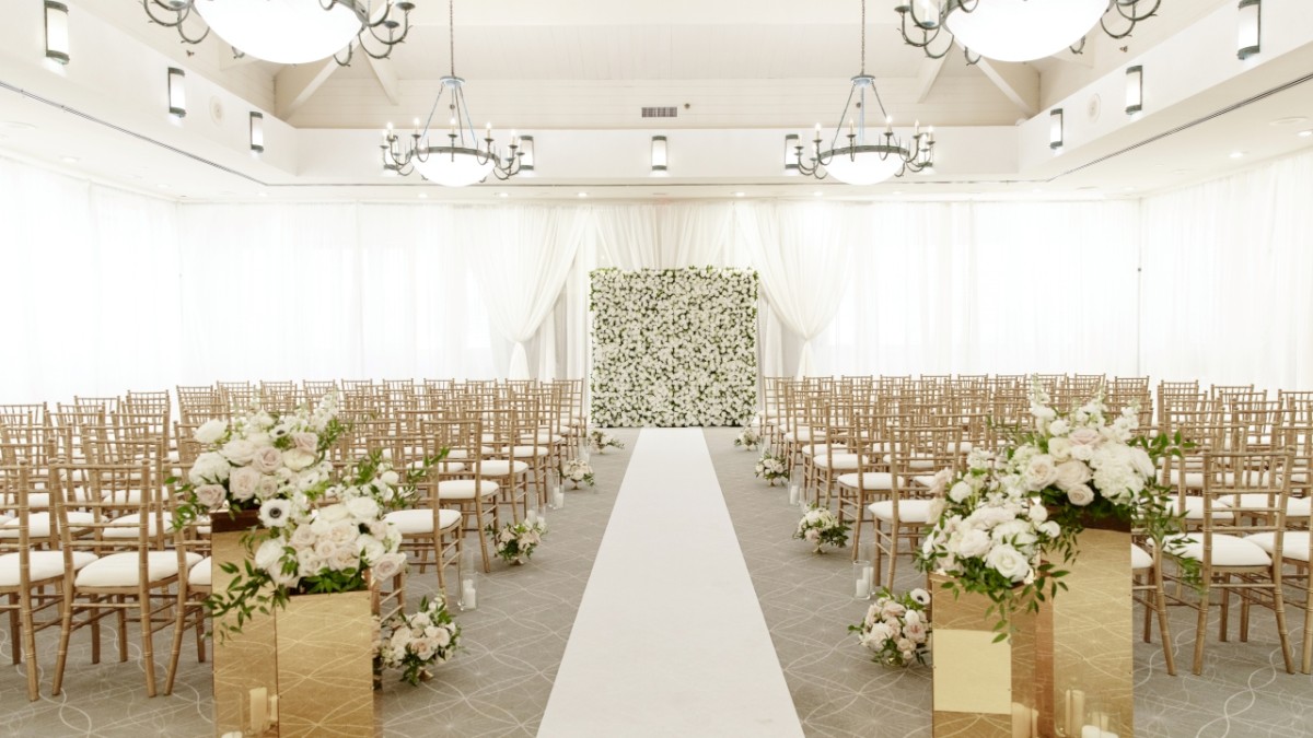 An elegantly decorated wedding venue with rows of chairs, floral arrangements, chandeliers, and an aisle leading to a flower-covered backdrop.