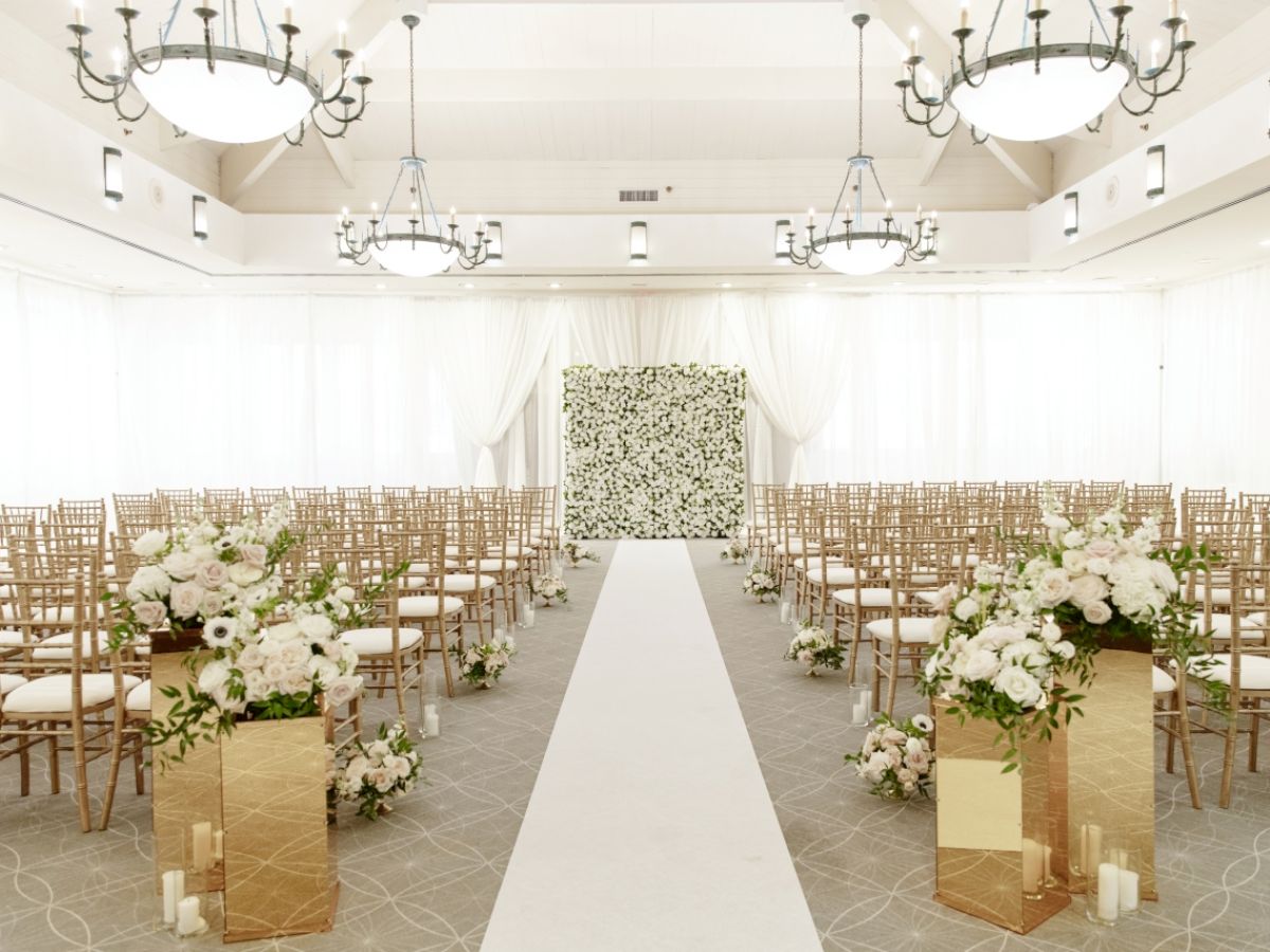 An elegantly decorated wedding venue with rows of chairs, floral arrangements, chandeliers, and an aisle leading to a flower-covered backdrop.