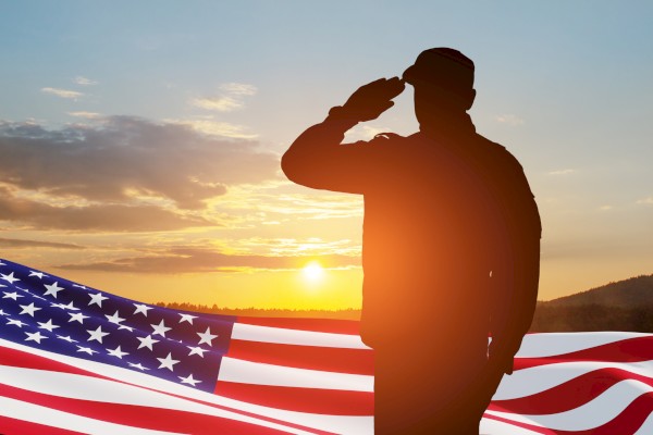 A silhouette of a person saluting against a sunset backdrop, with an American flag in the foreground. The scene evokes a patriotic moment.