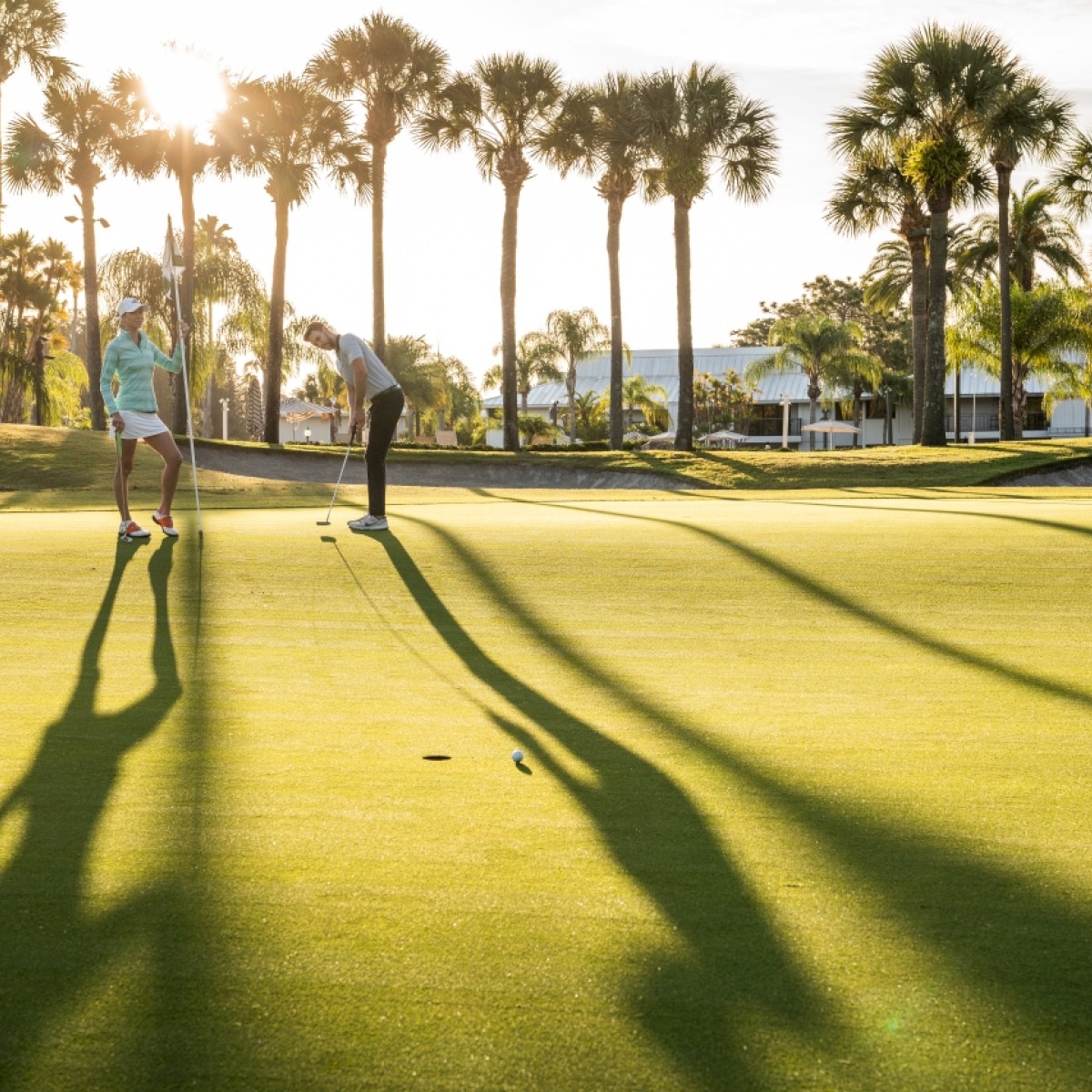 Two people on a golf course with palm trees casting long shadows at sunset.