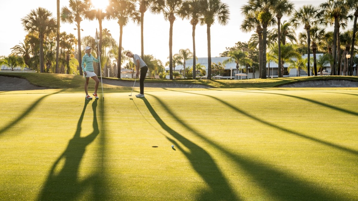 Two people on a golf course with palm trees casting long shadows at sunset.