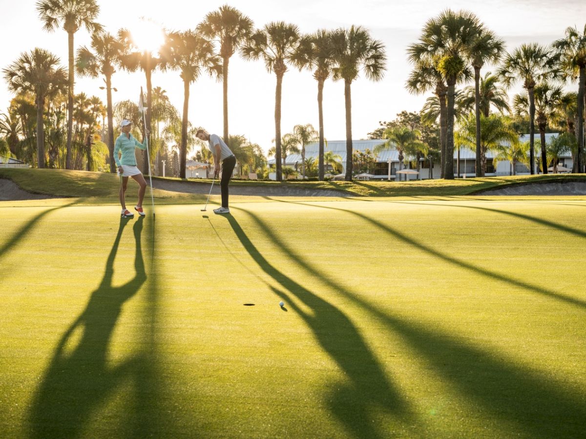 Two people on a golf course with palm trees casting long shadows at sunset.