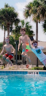 Four children wearing swimsuits and goggles are jumping into a pool simultaneously, with palm trees and a building in the background.