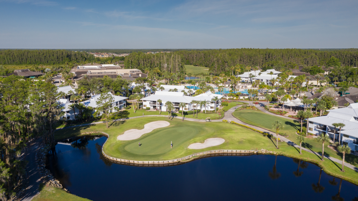 Aerial view of a golf course with sand traps, a pond, surrounding trees and clubhouse buildings in the background, under a blue sky ending the sentence.