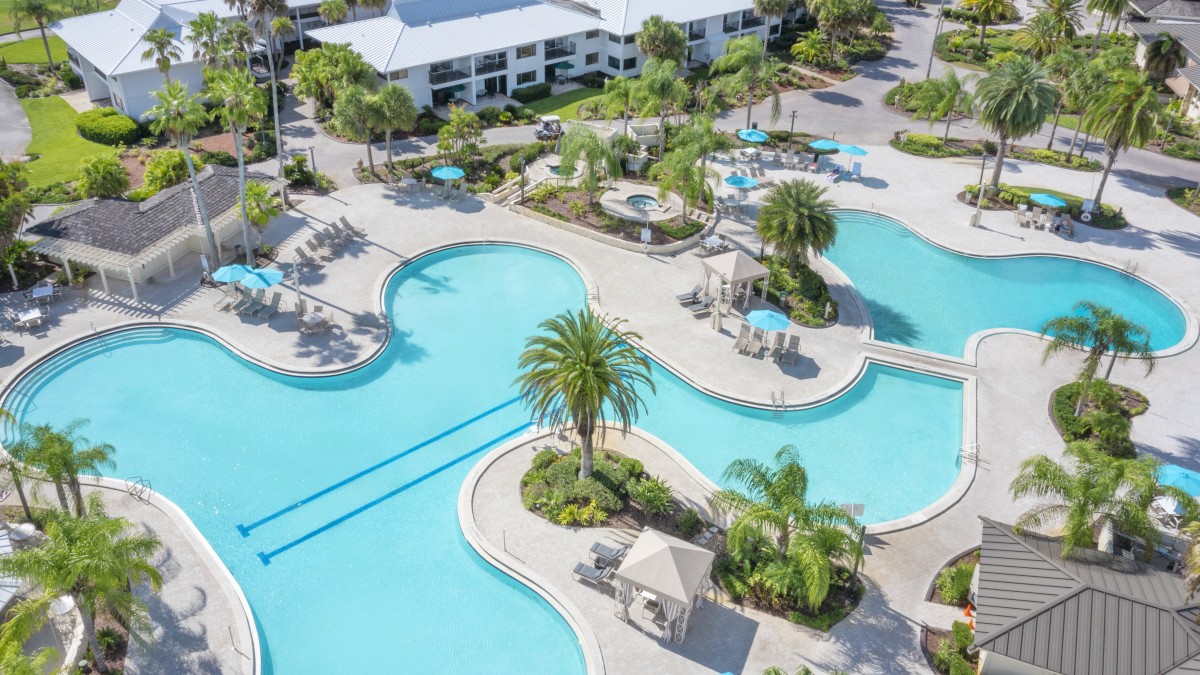 An aerial view of a large, resort-style swimming pool with surrounding lounge areas, palm trees, and nearby buildings.