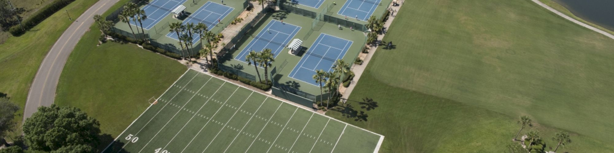 The image shows a sports complex with multiple tennis courts, a small football field, surrounding greenery, and a nearby body of water.