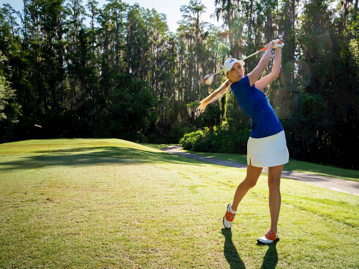 A woman in a blue shirt and white skirt is swinging a golf club on a lush, green golf course surrounded by trees.