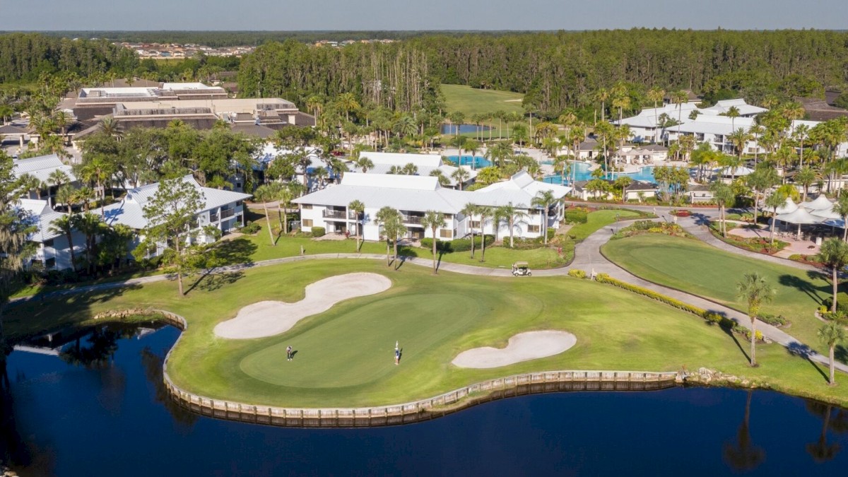 An aerial view of a golf course with several buildings and trees surrounding it, adjacent to a body of water, with a clear sky overhead.