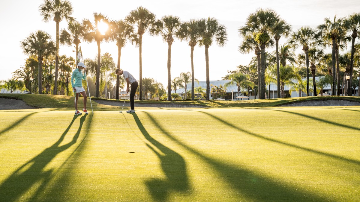 Two people are on a golf course under a sunny sky, surrounded by palm trees. Long shadows stretch across the green grass.