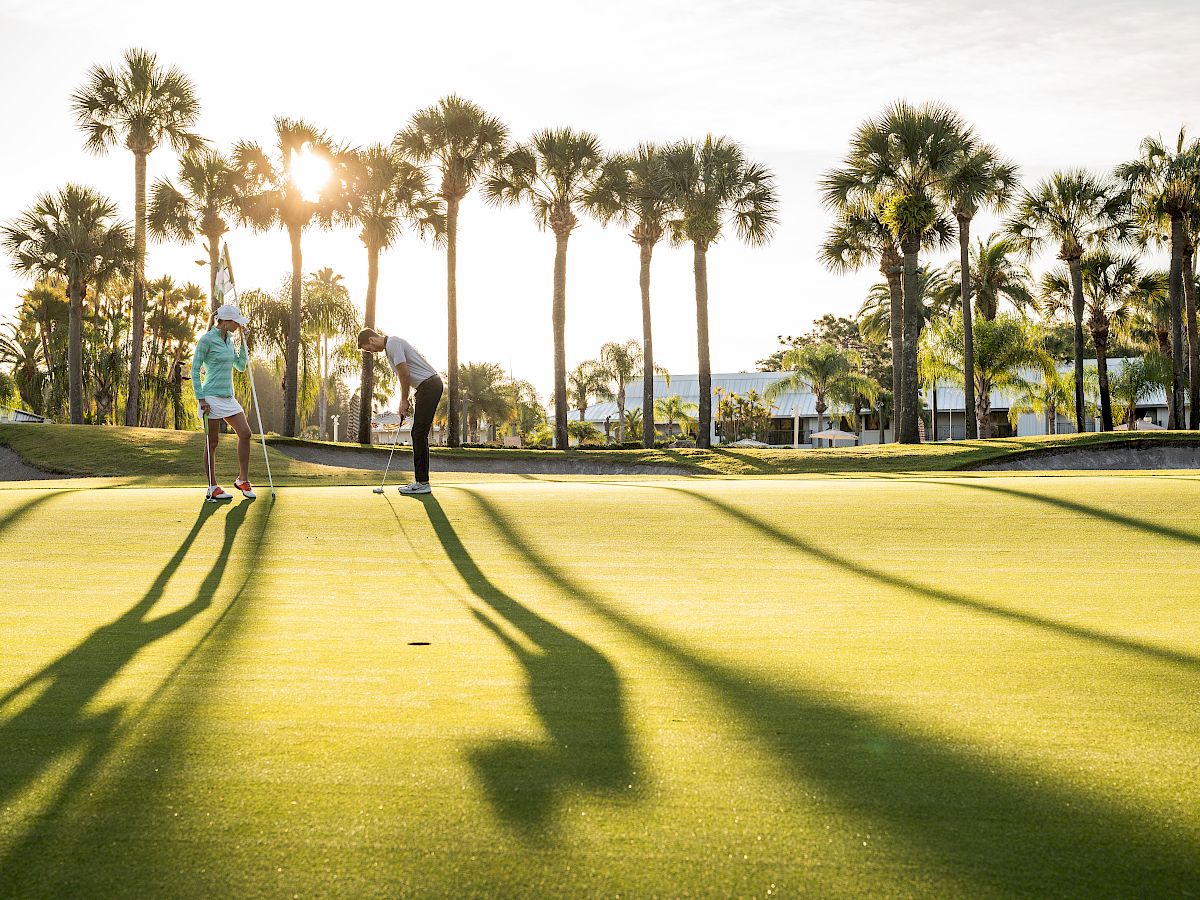 Two people are on a golf course under a sunny sky, surrounded by palm trees. Long shadows stretch across the green grass.