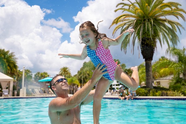 A man lifts a smiling child in a pool surrounded by palm trees and a blue sky, creating a joyful, sunny scene.