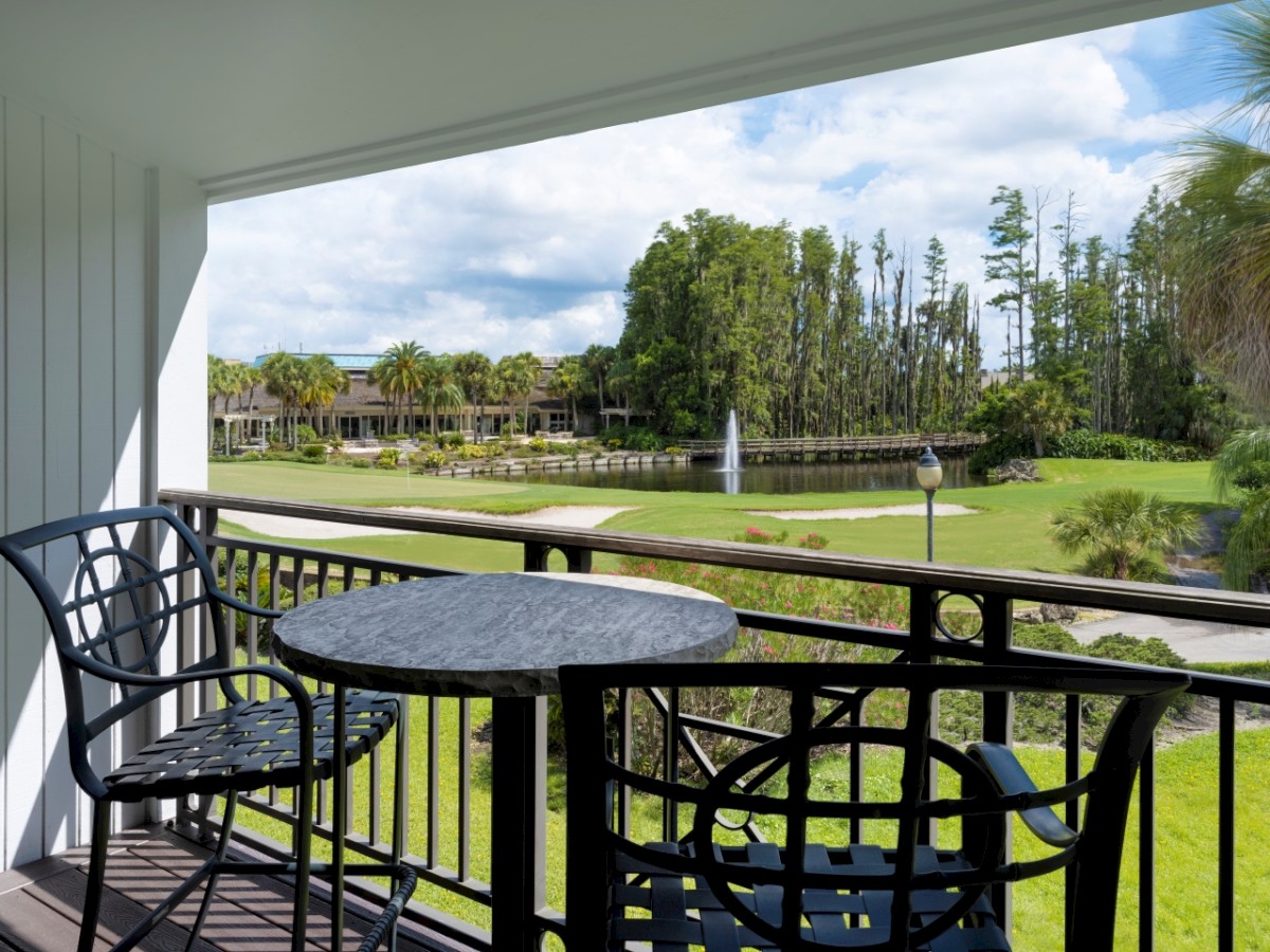 The image shows a balcony with a table and two chairs overlooking a well-maintained garden and a fountain, with trees and a cloudy sky in the background.