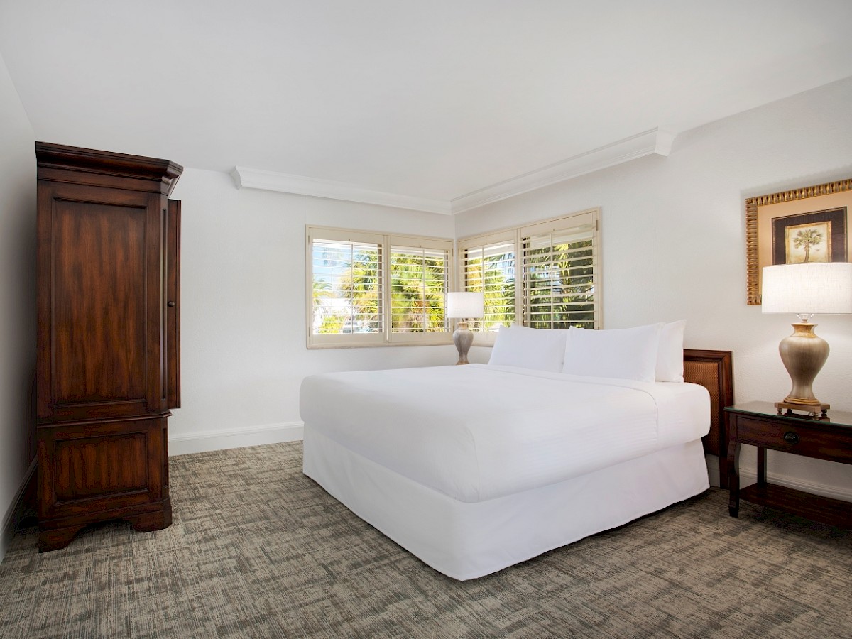 A clean, well-lit bedroom features a double bed, wooden armoire, side tables with lamps, and a window with shutters.