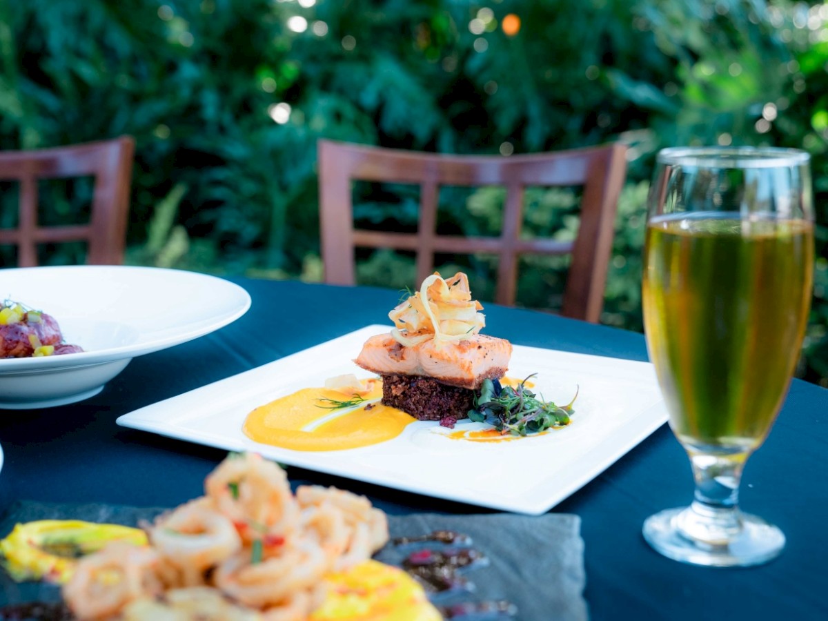 The image shows a table with a glass of beer, a dish with seafood, a side salad, and a plate of calamari, set in an outdoor dining area.