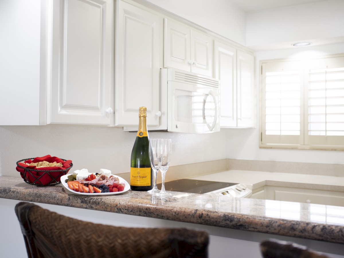 A kitchen with white cabinets and a granite countertop, featuring a bottle of champagne, two glasses, and a platter of snacks.