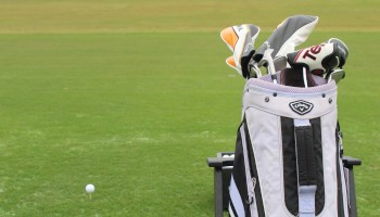 A golf bag with clubs stands on a golf course, near a golf ball, with a cloudy sky and grassy landscape in the background.