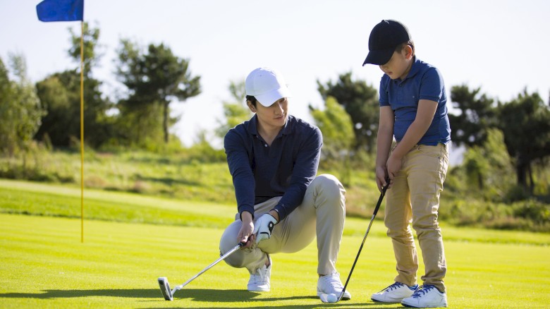 A person coaches a child in putting on a golf course, with a flag in the background and trees lining the horizon.