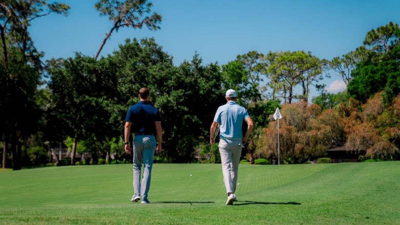 Two people walking on a golf course towards a flag, surrounded by trees and under a clear blue sky.