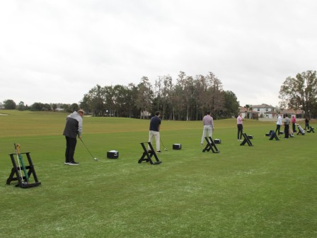 People practicing golf swings on a driving range, with equipment stands and a grassy field in the background.