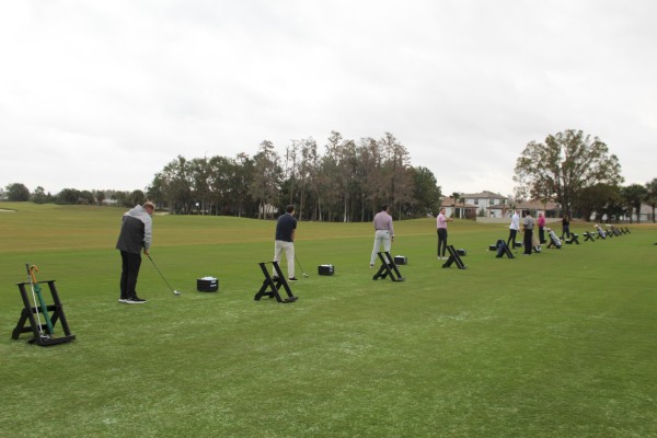 People practicing golf swings on a driving range, with equipment stands and a grassy field in the background.