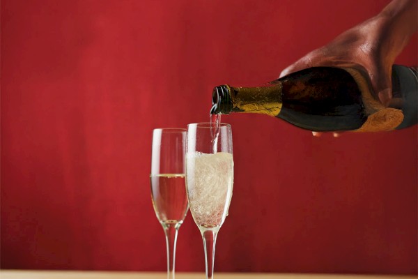 A hand pours champagne into a glass next to another full glass and two strawberries on a table, with a red background.