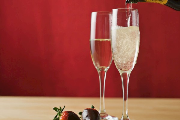 Two champagne flutes, one being filled, with chocolate-covered strawberries in front against a red background.