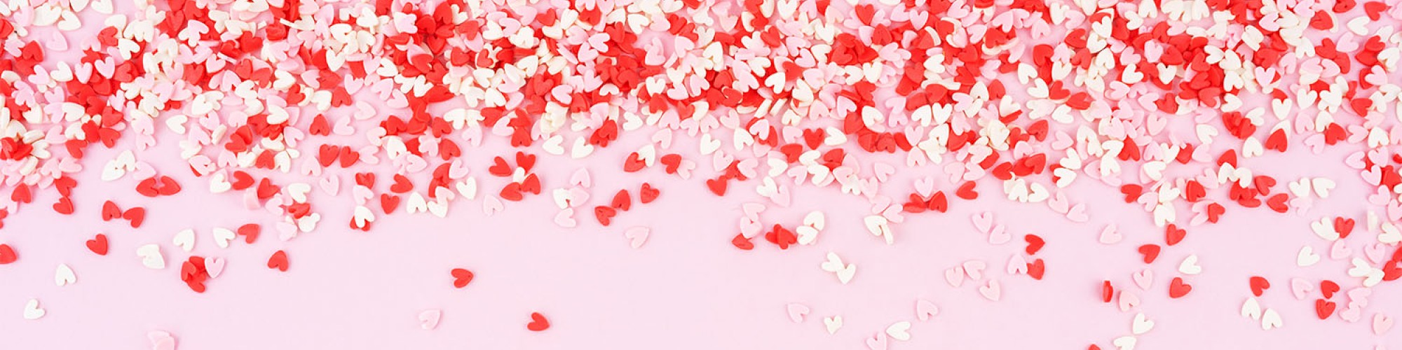 Red and white heart-shaped confetti scattered on a pink surface, concentrated at the top and gradually spreading out.