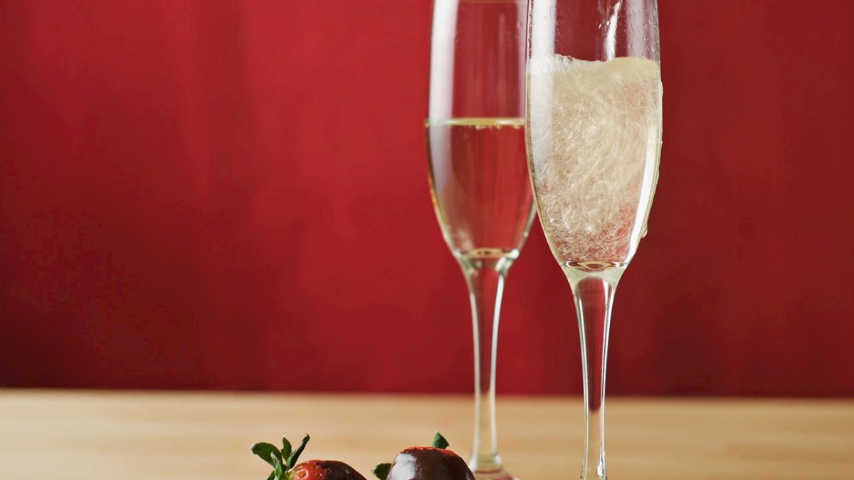 Two champagne flutes; one being filled, accompanied by chocolate-covered strawberries on a wooden surface against a red background.