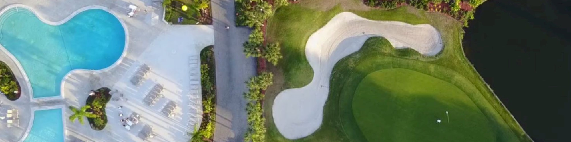 The image shows an aerial view of a resort featuring a swimming pool area on the left and a golf course with sand traps on the right, separated by a road.