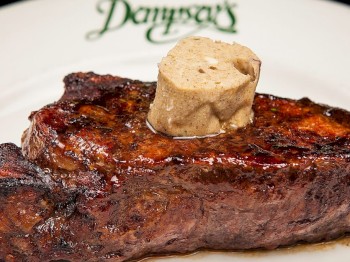 This image shows a grilled steak topped with a dollop of seasoned butter on a white plate with green script.