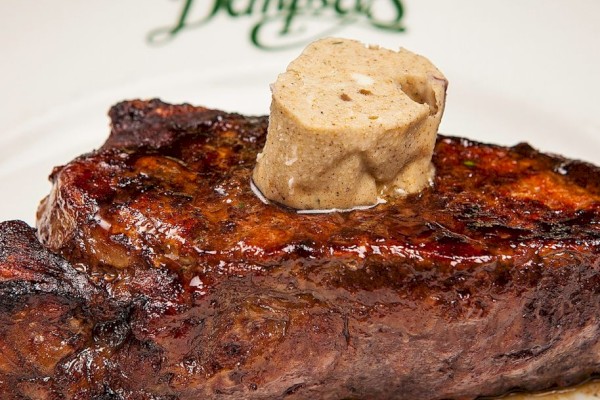 This image shows a grilled steak topped with a dollop of seasoned butter on a white plate with green script.