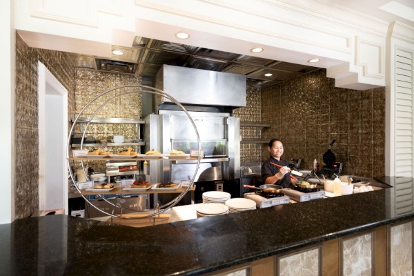 A chef is cooking in a stylish kitchen with a black countertop. The kitchen features stainless steel appliances and decorative tile walls.