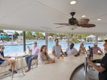People sitting at an outdoor bar near a pool, being served drinks by a bartender. It’s a sunny and pleasant environment, perfect for relaxing.