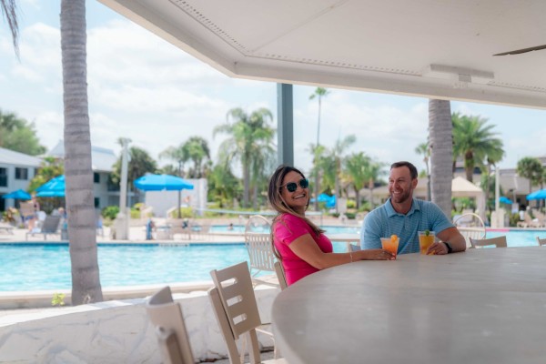 Two people are sitting at a poolside bar with drinks, enjoying the sunny weather.