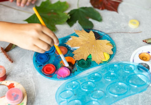 A hand holds a paintbrush near a palette with paint, surrounded by autumn leaves; ready for a painting activity.