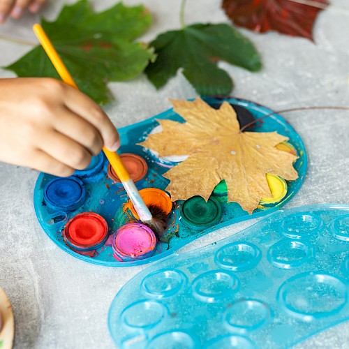 A hand holds a paintbrush near a palette with paint, surrounded by autumn leaves; ready for a painting activity.