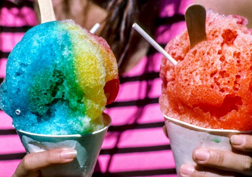 A person holds two colorful shaved ice treats in paper cups with wooden spoons, one blue and yellow, the other red, against a striped shirt background.