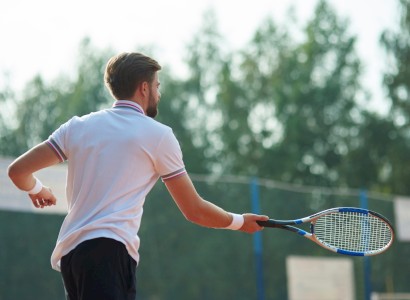 The image shows a man in a white shirt playing tennis on an outdoor court, holding a racket, and preparing to hit a ball.