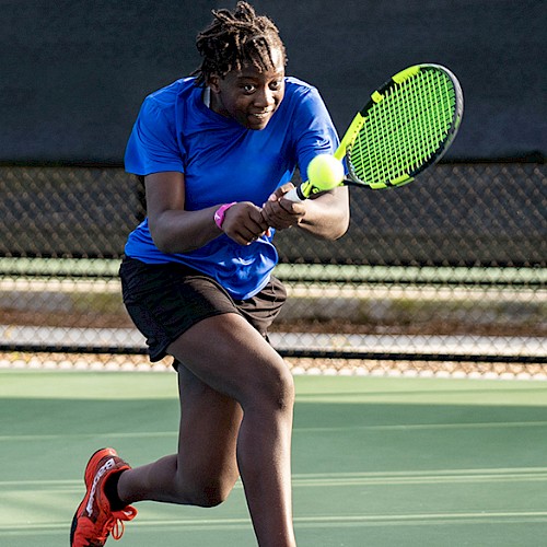 A person is playing tennis, taking a shot with a green racket. They are wearing a blue shirt, black shorts, and red shoes, on an outdoor court.