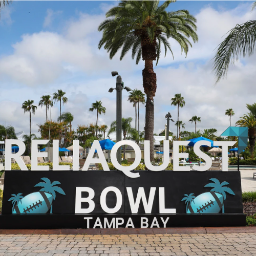 A sign for the ReliaQuest Bowl, held in Tampa Bay, is displayed against a backdrop of palm trees and a clear sky, with a tropical setting.