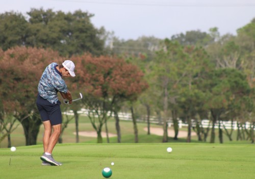 A golfer wearing a patterned shirt and shorts swings his club on a golf course, surrounded by trees.