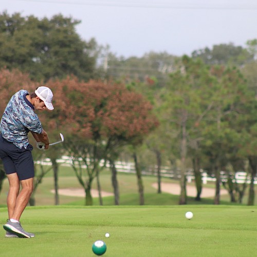 A golfer wearing a patterned shirt and shorts swings his club on a golf course, surrounded by trees.