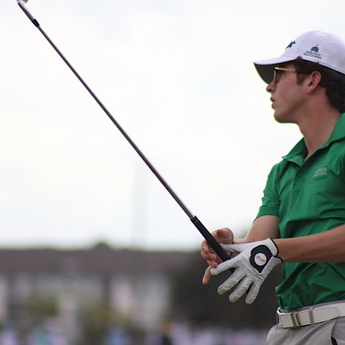 A person wearing sunglasses and a green shirt, holding a golf club likely after a swing. The background shows a blurred outdoor setting.
