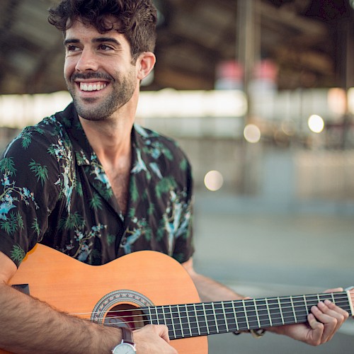 A smiling man playing a guitar, blurred background suggesting an indoor space.