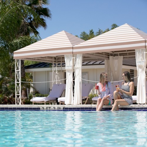Two people sit by a pool under striped cabanas, enjoying drinks and chatting on a sunny day, with palm trees in the background.