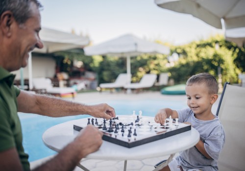 A man and a young boy are playing chess at a table by a poolside, enjoying a sunny day outdoors under some umbrellas, ending the sentence.