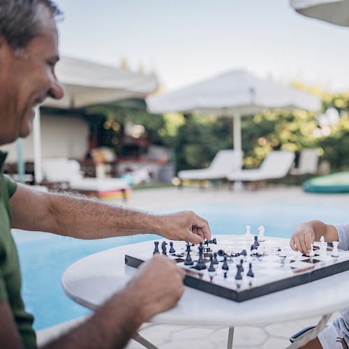 A man and a young boy are playing chess at a table by a poolside, enjoying a sunny day outdoors under some umbrellas, ending the sentence.